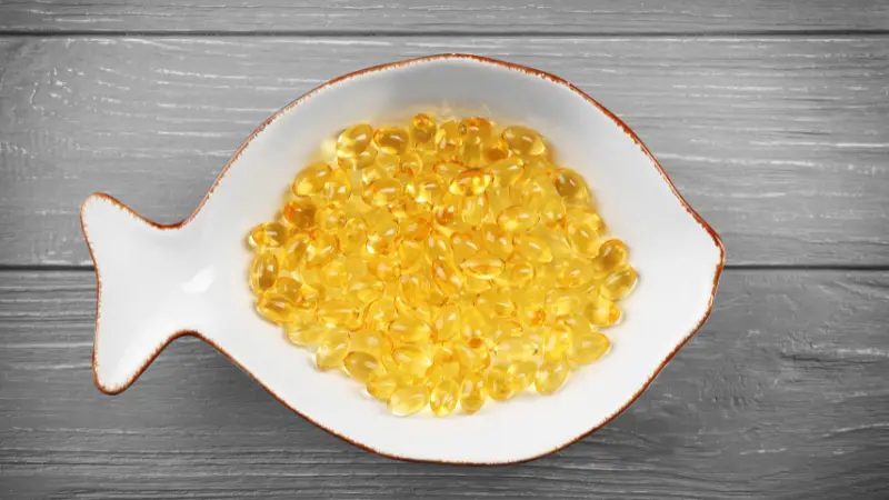 This image shows a fish in a bowl filled with golden oil beads. The fish is swimming in the oil and appears to be happy and content. The oil beads are shiny and reflect the light, making the image look bright and inviting. The wooden background adds a rustic and natural touch to the image.