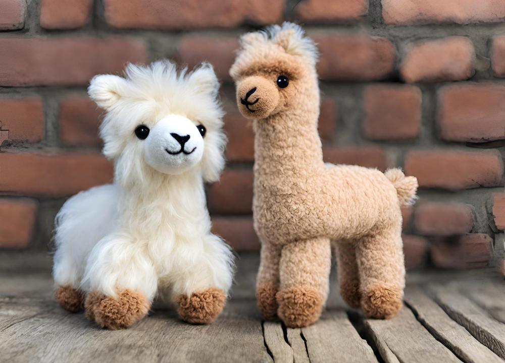 This image shows two stuffed alpaca dog toys, one brown and one white, sitting next to each other on a wooden surface in front of a brick wall.